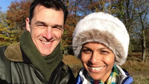 Thoulstone Park James and Alka Hughes-Hallett smile at the camera from outdoors, with trees behind them. It appears to be a cold day as They are in winter coats with scarves and Alka wears a fluffy hat