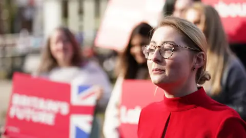 A woman in a red coat and glasses standing in front of a crowd of people holding signs supporting Labour. 