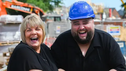 Allwyn Dannica and Adam Lopez standing outside at a building site. Dannica is a woman with a blonde bob who is looking at the camera and laughing, she is wearing a black T-shirt. Next to her, on the right, Adam Lopez is wearing a blue hard hat and is wearing a black T-shirt. He is looking directly at the camera and laughing.