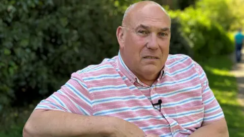 A man sits outside on a sunny day. He has a shaved head and a blue, red and white striped shirt. Behind him are green shrubs and grass.