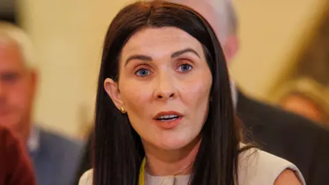 PA Media A woman is standing in front of a mic and talking to the media. There are other people standing around her. She has long brown hair and is wearing a yellow lanyard round her neck.
