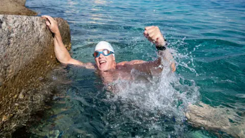 Lewis Pugh punches the air as he swims in the sea while wearing a white swim cap and blue goggles.