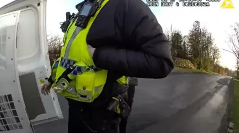 Footage from a body-camera shows a police officer, who is wearing a fluorescent yellow jacket, standing at the doors of a white police van.