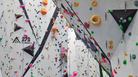 270 Climbing Park A view half way up one of the tall climbing walls at the centre. The white walls are sloped and jagged, dotted with various brightly coloured handles of different shaped and sizes for people to grab onto. There is a man wearing beige shorts, a pink top and a yellow harness climbing up the opposite wall.
