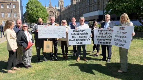 A group of people stand in front of the Houses of Parliament holding protest placards about the Cil levy in bright sunshine.