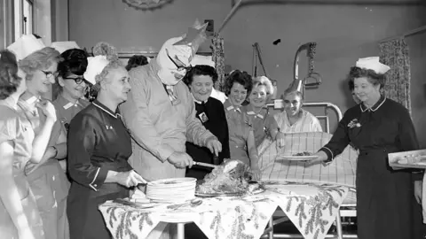 A black-and-white image of a man carving a turkey, surrounded by women in nurse's uniforms. The man is wearing overalls, bandages over his face and a star on his head as he carves the turkey on a table covered in a decorative festive cloth. A nurse holds out a plate for the turkey to be served. A male patient can be seen sitting up in a bed in the background, wearing a hat. All are smiling.