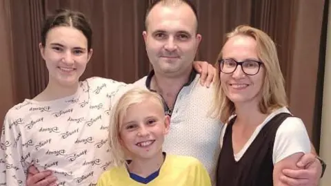 Family handout A family are standing in a landing of a house with brown curtains behind them. There is a man, woman, a girl and boy and they have their arms around each other smiling and wearing casual clothes.