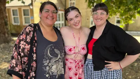 Stuart Woodward/BBC Three women standing next to each other.
(L-R) A woman wearing clear frame glasses and a oversized cardigan while smiling at the camera.
The woman next to her is wearing a pink co-rd that is light pink with red hearts on it.
The woman on the right is wearing a red crop-top paired with an oversized black cardigan and checked white and blue trousers. She has a fringe and is wearing clear framed glasses. She has one hand on her hip.
Behind them is trees and a building.