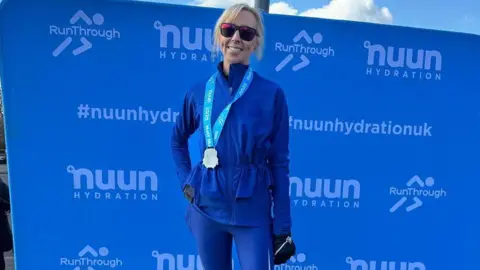 Supplied Danielle Everett stands in front of a blue banner which has sponsor and organiser logos on it. She has blond hair and is wearing sunglasses, a blue top and her medal around her neck.