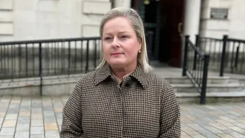 A woman with shoulder-length blonde hair wearing a brown and cream checked coat, stood outside the steps and railings of a courthouse