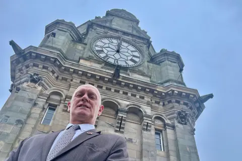 Brian Duncan looking down at the camera in front of the clock. There is a blue sky and he's wearing a grey suit and spotted tie.