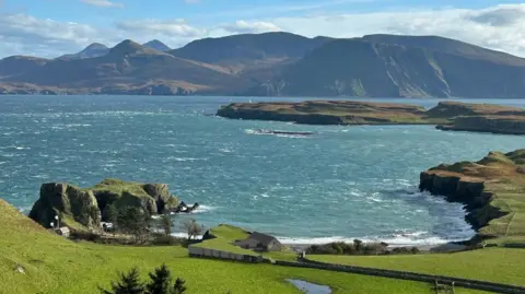A visualisation of a building in a curved bay with green fields in the foreground and mountains rising up across an area of sea