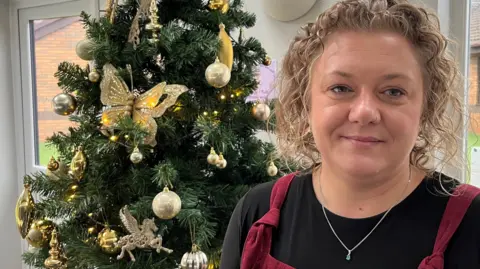 Cheryll Hamid who has curly blonde hair and wears a black top under some red overalls, stands in front of a Christmas tree