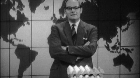 Cliff Michelmore standing in the studio. He's wearing a suit, has his arms crossed, and in front of him is a stack of dozens ofeggs.