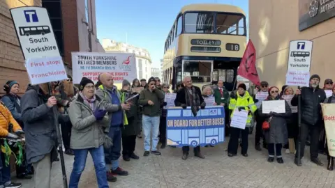 Spencer Stokes/BBC A group of 20-30 people holding cardboard signs with slogans about buses. In the background is a retro-looking disused beige bus.