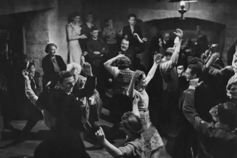 Getty Images A black and white image of the film's ceilidh scene. Men and women are dancing with their arms raised. The space they are in has stout stone walls. A group of people are standing by a wall watching the dancing.