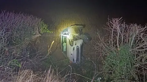 Nottinghamshire Police A crash white Fiat Punto on its side next to bushes with a front wheel hanging off