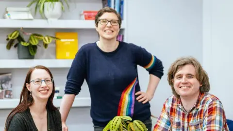 Birmingham Hippodrome/Angela Grabowska Jenni Pinnock, Helen Arney and Brian Mackenwells, smiling broadly, in a room with grey walls and shelves with books and plants on it. Jenni is seated left and has long reddish brown hair, is wearing glasses and a black top; Helen is standing in the middle and is wearing glasses and a blue jumper with a rainbow pattern; Brian is seated right and has light brown hair and is wearing a multi-colour plaid shirt. 