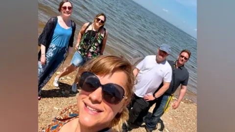 Toria Papworth A group of five people standing on a beach. They are all smiling at the camera with the sea behind them.