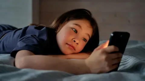 A young girl is seen lying on her front looking at a bright smartphone screen in a dark room 