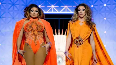 BBC/World of Wonder/Guy Levy Daniel Jervis pictured alongside drag queen Mariah Balenciaga. The pair are in full drag and wearing orange gowns.