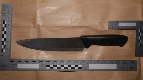 CPS A kitchen knife with a black handle resting on brown paper. Two rules show its height and width. 