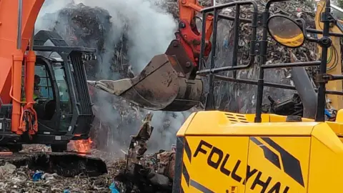 An excavator is being used to dig through a large pile of waste. Flames are seen in the background
