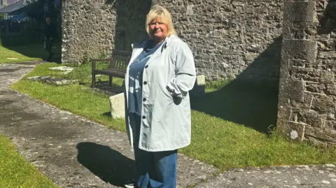 Kieran Molloy A woman with a blonde bob looking at the camera. She has a pale blue raincoat with a blue t-shirt and blue jeans. She is stood in front of a stone building and there is a stone path with grass on either side.