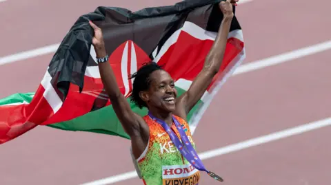 Faith Kipyegon, wearing an orange and green Kenyan athletics vest, with a gold medal on a purple ribbon bouncing around her neck, smiles broadly as she holds a Kenyan flag aloft behind her. The runner is visible from stomach up while a terracotta-coloured athletics track with white lines is seen out of focus in the background