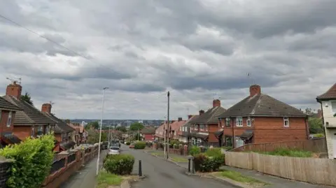 A street with several semi-detached houses on either side. There are street lights on the road and cars parked on the side. The sky is overcast. 