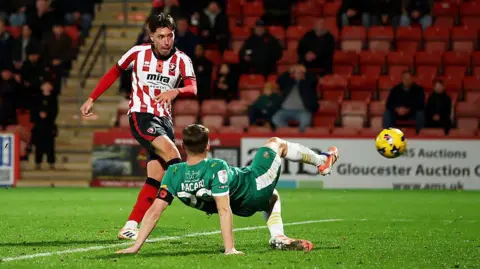 Isaac Hutchinson stands on one leg after striking the ball which flies through the air towards the goal, as a Notts County player crouches on the floor with a leg outstretched trying to block it