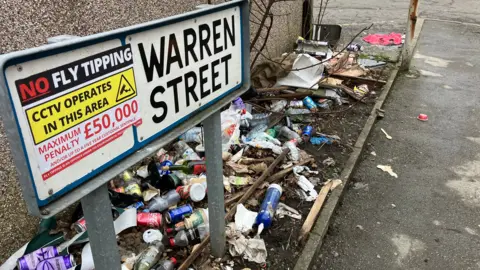 The Warren Street road sign has a sticker on it saying 'No Fly Tipping'. Beneath it are scores of empty cans and bottles. 