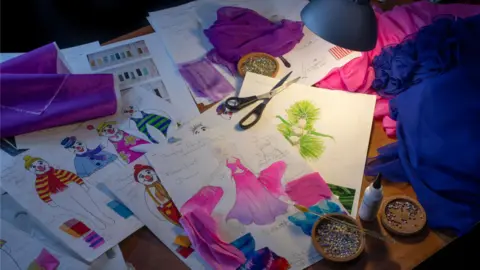 Thursford Christmas Spectacular Colourful paper sketches of costume designs are spread out on a wooden desk alongside materials, small bowls of pins and a pair of scissors.