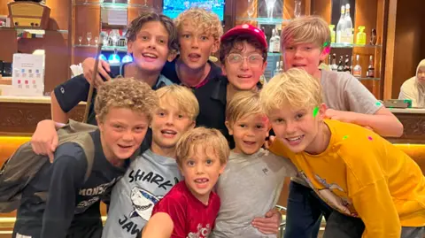 BBC A group of young boys in a small group smile at the camera. Many of them have long blonde hair and they are wearing T-shirt and shorts. 