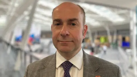 Professor Keith McLay is the Deputy Vice Chancellor at the University of Derby. He is looking directly into camera and is wearing a grey checked suit with a white shirt and purple tie.