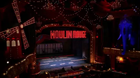 An empty stage of a theatre in the West End. The stage is lit up by lots of lights and has Moulin Rouge displayed in red above it. There is a large blue elephant to the right and a windmill to the left. 