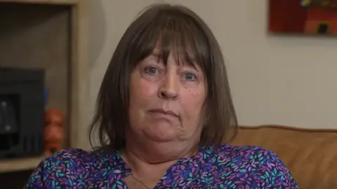 Whendie Backwell, a woman with blue eyes and shoulder-length brown hair, styled straight with a fringe. She is wearing a purple blouse. She is sitting on a brown sofa and has a blank expression on her face.
