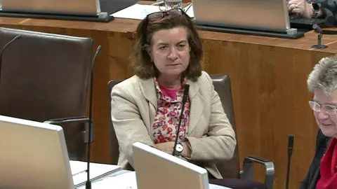 Eluned Morgan is pictured with her arms crossed wearing a white jacket sat in a chair in the debating chamber of the Welsh Parliament. Her colleague Jane Hutt  is sat next to her. They are listening to Andrew RT Davies, Welsh Conservative Senedd leader, who is not in the shot.