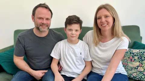 Rachel and Ralph sit on a green sofa with Henry between them. Ralph has short light brown hair, brown/grey stubble, and wears a grey t shirt with jeans. Henry has short brown hair and wears a white t shirt and dark bottoms. Rachel has long blonde hair and wears a white t shirt with jeans.