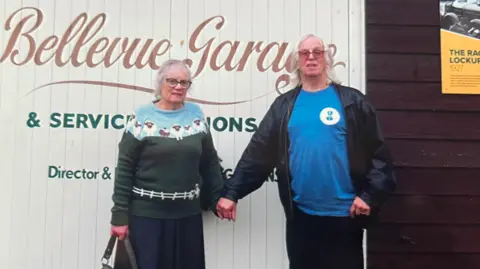 Worcestershire Acute Hospitals NHS Trust A woman in a green and blue jumper with sheep on it and a dark blue skirt is holding hands with a man in a blue top and black leather jacket. They are standing in front of shutters which read Bellevue Garage.