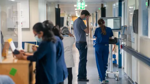 A view of a hospital corridor with various members of staff standing around - two look at a computer monitor