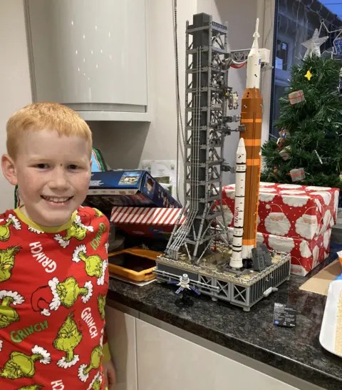 PA Media Efan standing beside his lego rocket on a kithen counter. He is wearing grinch pyjamas and is standing beside a mini christmas tree with wrapped presents. 
