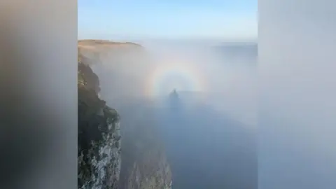 Chris Atkinson A Brocken Spectre - an image of a person's shadow captured in the mist. It has a circular rainbow surrounding it and the cliffs and sea can be seen in the image.