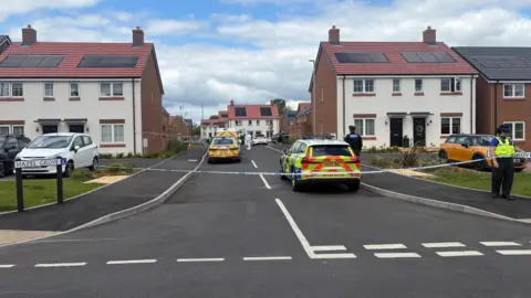The entrance to a housing estate viewed from a t-junction. Blue and white police tape has been stretched across the scene. Several police vehicles are parked on the road and a number of police officers are standing around.
