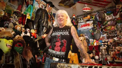 Martin leans against a red pool table surrounded by a dense collection of pop-culture memorabilia. The room contains masks, stuffed toys, posters, model displays and a life-sized figure dressed in black standing behind them.