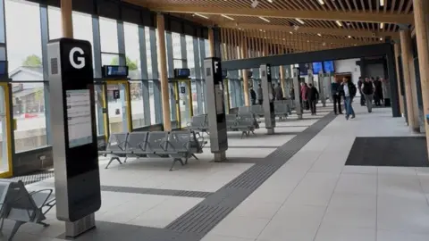 A row of bus stands inside the new Bishop Auckland bus station. There is seating and a digital screen at each stand. A group of people is walking down the hall.