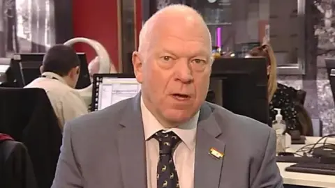 Stephen Giles-Medhurst wearing a grey jacket with a white shirt and dark grey tie which has a small white logo appearing several times. There is a media newsroom behind him.