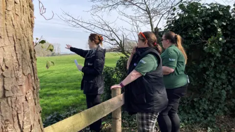 Three women are stood by a fence around a field. One of the women is raising her arm and gesturing towards the field as the other watch. 
