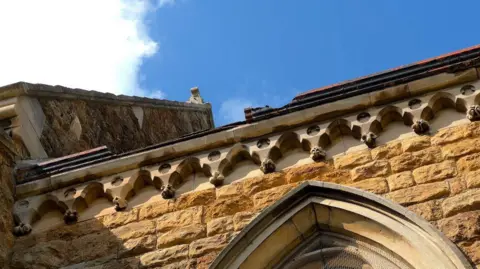 A section of the church which has been damaged by the cross. A horizontal metal piece of guttering has holes in it. 