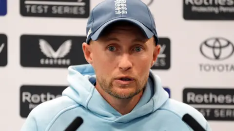 Joe Root speaks to the media with sponsors boards behind him